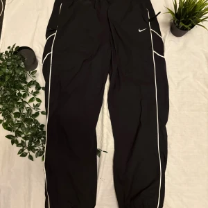 Svarta Nike byxor med vita detaljer - Svarta byxor från Nike med vita kontrastlinjer längs benen och broderad Swoosh-logga på framsidan. Byxorna har elastisk midja och avslappnad passform, perfekta för en sportig och chill stil. Materialet är lätt och känns skönt mot huden. Bra mot regn