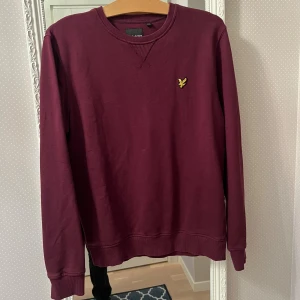 Vinröd sweatshirt från Lyle & Scott - Vinröd sweatshirt från Lyle & Scott med klassisk rund hals och gul broderad logga på bröstet. Tröjan är i mjuk bomull och har långa ärmar samt ribbade muddar vid ärmslut och nederkant. Perfekt för en chill och stilren look. Använd runt 5 gånger, skriv för mer bilder och info! Inga fläckar eller liknande.