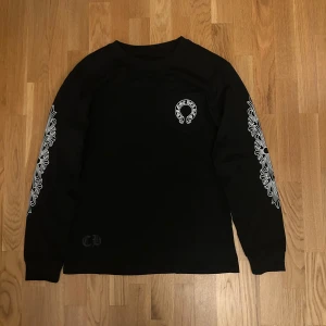 Svart långärmad Chrome Hearts tröja - Svart långärmad tröja från Chrome Hearts med stora vita tryck på ryggen och bröstet samt detaljerade vita mönster längs båda ärmarna. Tröjan har rund hals och är i mjuk bomull, perfekt för dig som gillar streetwear med attityd.