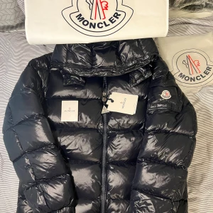 Moncler Maya navy blue  - Snygg mörkblå pufferjacka från Moncler med glansig finish och klassisk logga på ärmen. Jackan har dragkedja framtill, hög krage och quiltad design. Insidan är vinröd med ett unikt serietryck och märkning för äkthet. Perfekt för kalla dagar när du vill hålla stilen. Storlek 5/xl 0bs lnte 0rglnal!!