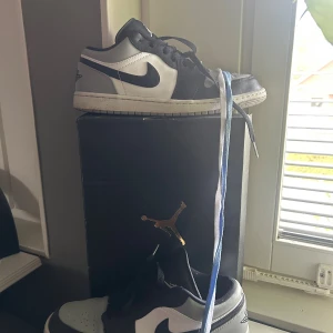 Nike Air Jordan 1 Low svart/vit - Säljer ett par Nike Air Jordan 1 Low i svart, vitt och grått skinn. Klassisk siluett med svart swoosh, perforerad tå och snörning. Perfekta för dig som gillar streetwear och vill ha en ikonisk sneaker i garderoben. Vitt o blåa extra snörren till kommer.