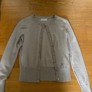 Beige kofta från Holly & Whyte XS - Snygg beige kofta från Holly & Whyte i storlek XS. Klassisk modell med rund halsringning, ribbade muddar och knappar framtill. Perfekt att slänga över en topp när det blir lite kyligt. Mjuk och skön känsla.