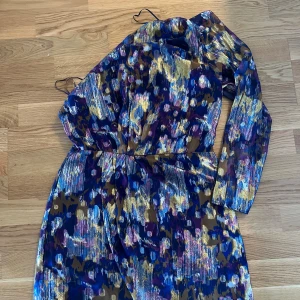Glittrig långklänning i blått och guld - Säljer en glittrig långklänning med abstrakt mönster i blått, guld, lila och inslag av rosa. Klänningen har långa ärmar och en figurnära passform i midjan. Materialet skimrar snyggt och ger en festlig känsla. Perfekt för dig som vill sticka ut med färg och glitter!