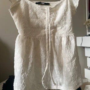 Vit blus med spets från BikBok - Supersöt vit blus från BikBok med broderad spets och knytband framtill.Tröjan har korta ärmar och en lätt volang vid axlarna. Perfekt för alla tillfällen och har en romantisk vibe med sin strukturerade spets och feminina siluett.