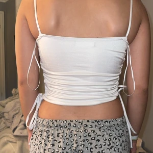 Vit croppad linnetopp med snörning - Säljer en vit croppad linnetopp med tunna axelband och snygg snörning i sidorna. Toppen har en tight passform och är tillverkad i ett glansigt, stretchigt syntetmaterial. Perfekt för dig som gillar trendiga och stilrena plagg med det lilla extra.