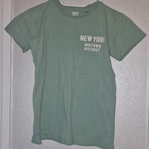 Grön t-shirt från lager 157 med tryck - Snygg grön t-shirt från lager 157 i 100% bomull med vitt tryck på bröstet där det står 'NEW YORK MIDTOWN DISTRICT'. Klassisk rund hals och korta ärmar, perfekt för en avslappnad stil. Det är lite ytludd på tröjan och en diskret fläck som syns på tröjans nederkant (se bild).