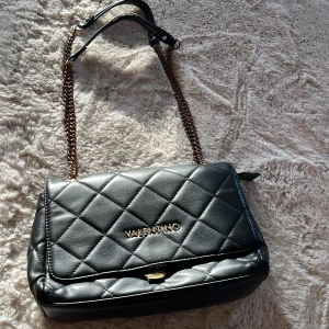 Svart quiltad axelväska Valentino - Snygg svart axelväska från Valentino i quiltat skinn med guldfärgade kedjor och detaljer. Väskan har lock med magnetlås, dragkedja under locket och ett klassiskt rutmönster. Perfekt storlek för det viktigaste och ger en lyxig känsla till din outfit.  Bra skick förutom att bandet har blivit rosé guld, detta ser ut att vara naturligt och att det ska vara den färgen. Sedan en liten bit gått upp där vid spännet syns på bilderna på väskan. Skicka för fler detaljer eller frågor 💕
