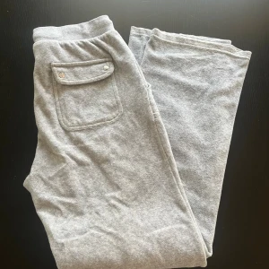 Grå Juciy Counture mjukisbyxor med ficka - Grå mjukisbyxor i bomull med elastisk midja och en ficka med lock och knapp bak. Perfekta för chill dagar eller när du vill vara extra bekväm. Klassisk rak passform och mjukt material. Storlek 14/15 år