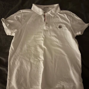 Vit Moncler piké med krage - Vit pikétröja från Moncler med klassisk krage och knappslå med röd, vit och blå detalj. Broderad Moncler-logga på bröstet. Kortärmad och tillverkad i mjuk bomull, perfekt för en clean och stilren look. I använt men bra skick. Storlek M men skulle säga att de passar som en S. Såklart helt äkta
