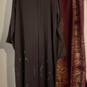 Mörkbrun långklänning med paljettdetaljer - Säljer en mörkbrun långklänning med raka, vida ärmar och diskreta svarta paljettdetaljer i vågiga mönster längs klänningen. Klänningen har en enkel, rak passform och är tillverkad i ett mjukt, lätt material som faller snyggt.Ganska lång för nån som är 1,65 neråt passar lite längre tjejer🫶🏾