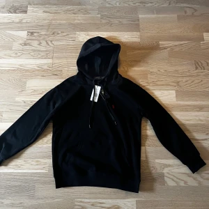 Svart hoodie från Polo Ralph Lauren - Svart hoodie från Polo Ralph Lauren med klassisk huva och dragsko. Tröjan har en liten röd broderad logga på bröstet, känguruficka och ribbade muddar. Tillverkad i mjuk bomull för en skön och avslappnad stil. Dock är prislappen av klippt men man får med den endå tvättlappen också för den skavde. Perfekt nu när de börjar bli lite kyligare . OBS liten i storleken