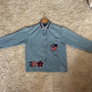 Jeansskjorta med broderier från Three Rivers - Ljusblå jeansskjorta från Three Rivers med unika broderier i rött, blått och beige. Skjortan har lång ärm, knappar framtill och coola detaljer som stjärnor och hjärtan på krage, bröstficka och nedtill. Perfekt för dig som gillar statement-plagg och denim.  (Jag som är med på bilderna är 190cm lång och väger 78kg)