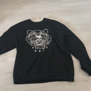 Svart sweatshirt med tiger från Kenzo - Svart sweatshirt från Kenzo med ikoniskt broderat tigerhuvud och texten 'Kenzo Paris' framtill. Tröjan har rund hals, långa ärmar och är tillverkad i mjuk bomull. Perfekt för dig som vill sticka ut med en streetig vibe.