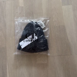 Svart ribbad mössa från C.P. Company - Svart ribbstickad mössa från C.P. Company med deras ikoniska goggles framtill. Tillverkad i mjukt material och perfekt för dig som vill sticka ut med en streetwear-inspirerad look. Mössan har en klassisk form och tydlig logga på glasögonen.