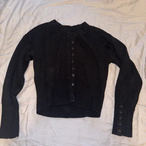 Svart stickad kofta med knappar - Svart stickad kofta med långa ärmar och knappar framtill. Modellen är croppad och har ribbade muddar samt extra knappar vid ärmsluten. 