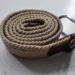 Beige flätat bälte med skinn-detaljer - Snyggt beige bälte med flätad design och bruna skinn-detaljer vid spännet. Bältet har ett klassiskt silverfärgat metallspänne och passar perfekt till jeans eller chinos. Den grova flätningen ger en cool och avslappnad vibe.