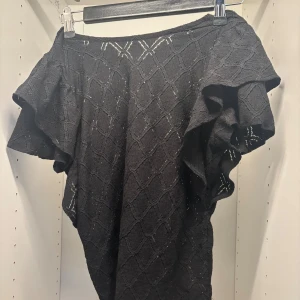 Svart blus med volangärm Holly & Whyte - Svart blus från Holly & Whyte med snyggt strukturmönster och volangiga korta ärmar. Toppen har en lätt croppad passform och diskret rutmönster i tyget. Perfekt för dig som gillar en stilren men ändå unik look.