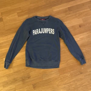 Blå sweatshirt från Parajumpers - Säljer en blå sweatshirt från Parajumpers med stort vitt tryck på bröstet och klassisk rund hals. Tröjan har långa ärmar och ribbade muddar samt en snygg patch på ena ärmen. Perfekt för dig som gillar streetwear och vill ha något stilrent. Storlek är L i Junior storlek skulle säga att den är S-M i vanlig storlek. Är i mycket bra skick bara använt ett par gånger. Säljer då den ej passar mig längre. Kontakta vid fler bilder eller funderingar.