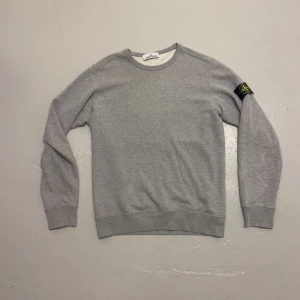 Grå sweatshirt från Stone Island - Säljer en grå sweatshirt från Stone Island med klassisk patch på vänster ärm. Tröjan har rund halsringning och långa ärmar. Tillverkad i mjuk bomull.