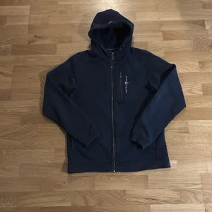 Marinblå hoodie från Sail Racing - Säljer en marinblå hoodie från Sail Racing med dragkedja och huva. Tröjan har en bröstficka med dragkedja och är tillverkad i en mjuk bomulls- och polyestermix. Perfekt för dig som gillar sportig och stilren look.    176=S