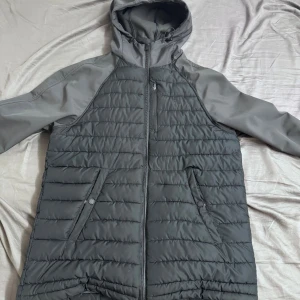 Grå quiltad jacka från Jack Wills - Säljer en grå quiltad jacka från Jack Wills med huva och dragkedja. Jackan har två snedställda fickor framtill och är tillverkad i polyester. Perfekt för kyliga dagar med sin lätta och smidiga design.