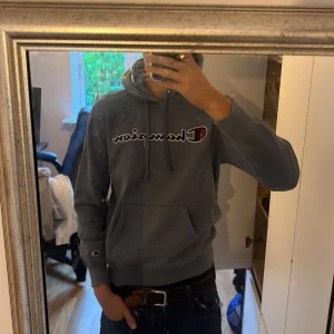Grå Champion hoodie med logga - Säljer en grå hoodie från Champion i storlek small. Tröjan har stor broderad Champion-logga på bröstet, snörning i huvan och en klassisk känguruficka framtill. Mjuk insida och ribbade muddar vid ärmslut och nederkant.