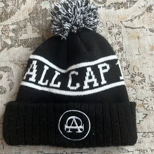 Svart och vit mössa med tofs från All Cap 1 - Svart stickad mössa med vit text 'ALL CAP 1' och broderad logga framtill. Mössan har en stor svartvit tofs på toppen och vita ränder runt om. Perfekt för kalla dagar och snygg streetstil.