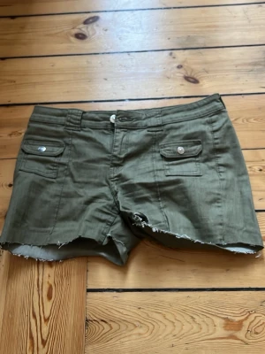 Olivgröna shorts med rå kant - Snygga olivgröna shorts med rå, fransig kant och två fickor framtill med knappdetaljer. Klassisk midja och bälteshällor. Perfekta för varma dagar och en avslappnad stil.