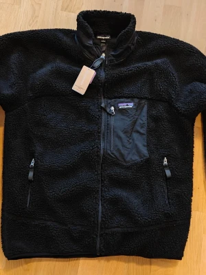 Patagonia Retro-X jacket, oanvänd - Helt ny och oanvänd, lite för liten. Är storlek XL enligt etikett men tycker helt klart den sitter mer som en L. Väldigt liten i storleken, ha detta i åtanke. Mysig svart teddyjacka från Patagonia med hög krage och hel dragkedja. Jackan har en bröstficka med dragkedja och två sidofickor med dragkedja. Tillverkad i fluffigt teddy-material som håller dig varm. Klassisk Patagonia-logga på bröstet och detaljer i svart tyg.