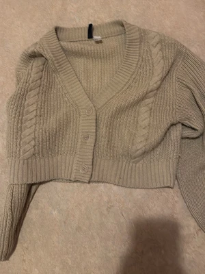 Beige cropped stickad kofta - Snygg beige cropped kofta med kabelstickade detaljer längs sidorna och bred ribbad kant. Koftan har djup v-ringning, knäppning framtill med tre knappar och långa ärmar. Perfekt att slänga över en topp för en chill look.