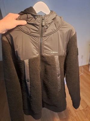 Svart teddyjacka från RevolutionRace - Mysig svart teddyjacka från RevolutionRace med huva och dragkedja framtill. Jackan har släta partier på bröstet och axlarna samt två vertikala fickor med dragkedja. Materialmix av fluffig teddy och slät polyester ger en sportig look.