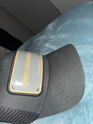 Svart truckerkeps John Hatter & Co - Säljer en svart truckerkeps från John Hatter & Co med en stor metallskylt framtill där det står 'CINDER-FUCKIN-ELLA'. Kepsen har mesh på sidorna och bak för extra ventilation och en böjd skärm. Perfekt för dig som vill sticka ut med en edgy accessoar.