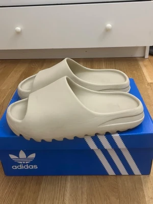 Beige Yeezy Slides - Säljer ett par stilrena beige tofflor från Adidas. De har en chunky sula med räfflat mönster och öppen tå. Tillverkade i ett mjukt och flexibelt material som ger skön komfort. Perfekta för chill dagar hemma eller till och från träningen.