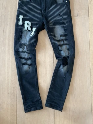 Svarta distressed jeans från amiri - Säljer ett par svarta slim fit jeans med slitningar och patchade bokstäver framtill och baktill. Jeansen har en tvättad look, råa detaljer och broderade bokstäver i vitt och grått. Perfekta för dig som gillar streetwear och unik stil.