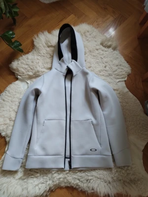 Ljusgrå hoodie från Oakley - Snygg ljusgrå hoodie från Oakley med svart dragkedja och svart kant på insidan av huvan. Hoodien har stora fickor framtill och Oakleys logga tryckt på ena fickan. Perfekt för en avslappnad och sportig stil. Är en rätt så liten M. Några små rivmärken på ena ärmen men annars i mycket bra skick