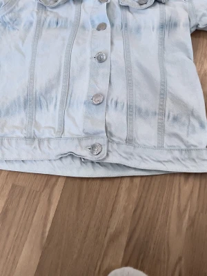Ljusblå jeansjacka med fickor - Snygg ljusblå jeansjacka med klassisk knappstängning framtill och två bröstfickor med lock. Jackan har en rak passform och tydliga sömmar som ger en cool vintagekänsla. Perfekt att slänga över en hoodie eller t-shirt.