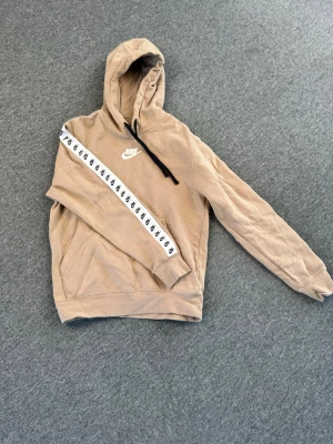 Beige Nike hoodie - Snygg beige hoodie från Nike med huva och svart dragsko. På vänster ärm finns ett vitt band med svarta Nike-loggor som ger en sportig vibe. Klassisk känguruficka framtill och Nike Swoosh broderad på bröstet. Perfekt för chill dagar eller streetwear-stil.
