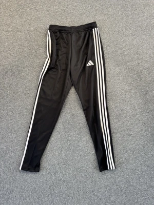 Svarta Adidas träningsbyxor - Säljer ett par svarta träningsbyxor från Adidas med klassiska vita ränder längs sidorna och Adidas-logga på låret. Byxorna har smal passform, resår i midjan och är gjorda i ett glansigt, snabbtorkande material som passar perfekt till träning eller chill.