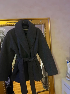 Mörkgrå kappa från Gina Tricot - Snygg mörkgrå kappa från Gina Tricot i premiumkvalitet. Modellen har bred sjalkrage, knytskärp i midjan och raka ärmar. Perfekt för dig som gillar en stilren och trendig look. Materialet känns mjukt och värmande, passar bra till lager-på-lager.