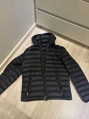 Svart dunjacka från Polo Ralph Lauren - Svart dunjacka med huva från Polo Ralph Lauren. Jackan har två dragkedjefickor framtill och klassisk quiltad design. Märkets röda logga är broderad på bröstet. Perfekt för kalla vinterdagar och riktigt snygg till streetwear.