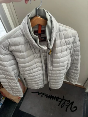 Ljusgrå dunjacka Parajumpers L - Snygg ljusgrå dunjacka från Parajumpers i storlek L. Jackan är superlätt, har quiltad design, hög krage och dragkedja framtill. Detaljer med orange och gul rem vid halsen ger en cool touch. Perfekt för kalla vinterdag och hösten och våren. Kvittot syns på bilderna och vid mer frågor hör av er.