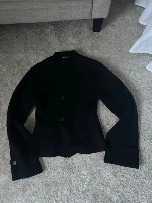 Svart tröja från Gina Tricot - Stilren svart tröja/kavaj från Gina Tricot med två knappar framtill och breda ärmslut med knappdetalj. Tröjan har en rak passform och är perfekt för en trendig look. Materialet känns mjukt och bekvämt, strl M men sitter mer som en S. Säljer för 200kr