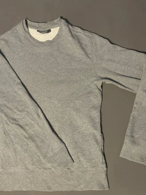 Grå sweatshirt från J.Lindeberg - Klassisk grå sweatshirt från J.Lindeberg med diskret logga broderad på bröstet. Tröjan har rund halsringning, långa ärmar och ribbade muddar vid ärmslut och nederkant. Tillverkad i mjukt bomullsmaterial för en avslappnad look.