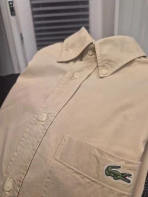 Beige skjorta från Lacoste storlek 38 - Klassisk beige skjorta från Lacoste med lång ärm, knappar framtill och bröstficka med den ikoniska gröna krokodilloggan. Skjortan har en avslappnad passform och är tillverkad i mjuk bomull. Perfekt för en clean och stilren look.
