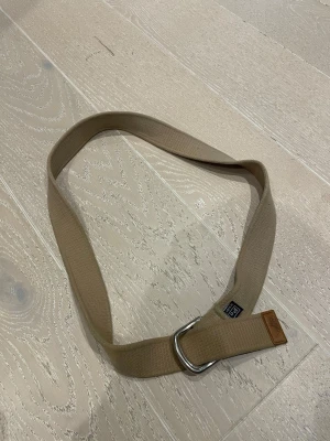 Beige Polo bälte med metallspänne - Snyggt beige bälte från Polo Ralph Lauren med dubbla D-ringspännen i metall och en detalj i brunt läder vid änden. Bältet är gjort i ett slitstarkt tygmaterial och har en klassisk, enkel design som passar till det mesta. Org pris: 800