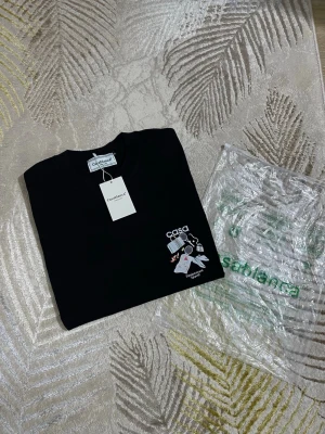 Svart t-shirt från Casablanca - Snygg svart t-shirt från Casablanca med tryck på bröstet och rund hals. T-shirten har en cool grafisk detalj med texten 'casa' och illustration. Tillverkad i mjuk bomull som känns skön mot huden. Perfekt för dig som gillar streetwear och trendiga plagg.