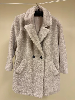 Beige teddykappa med dubbelknäppning från H&M - Mysig beige teddyjacka med bred krage och dubbelknäppning framtill från H&M Divided. Kappan har två snedställda fickor och är fodrad på insidan. Perfekt för kalla dagar när du vill vara både varm och snygg. Materialet är fluffigt och ger en trendig look. Kappan är stor, större än XL den är märkt med. Omkretsen under armarna är 140 cm och den är 88 cm lång. 