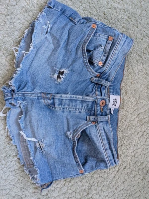 Blå jeansshorts från 157 med slitningar - Säljer ett par ljusblå jeansshorts från 157 med råa kanter och slitna detaljer. Shortsen har fem fickor, knappgylf och klassisk denimlook. Perfekta för varma dagar och ger en avslappnad vibe.