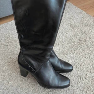 Svarta höga läderstövlar med klack - Snygga svarta stövlar i äkta läder med hög klack och dekorativa nitar vid ankeln. Stövlarna har spetsig tå och diskreta sömmar längs skaftet. Perfekta för att lyfta vilken outfit som helst.