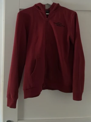 Vinröd hoodie från Peak Performance - Snygg vinröd hoodie från Peak Performance med dragkedja framtill och två fickor. Klassisk huva och diskret logga på bröstet. Tillverkad i mjukt bomullsmaterial som känns skönt mot huden. Perfekt för en avslappnad och sportig stil.